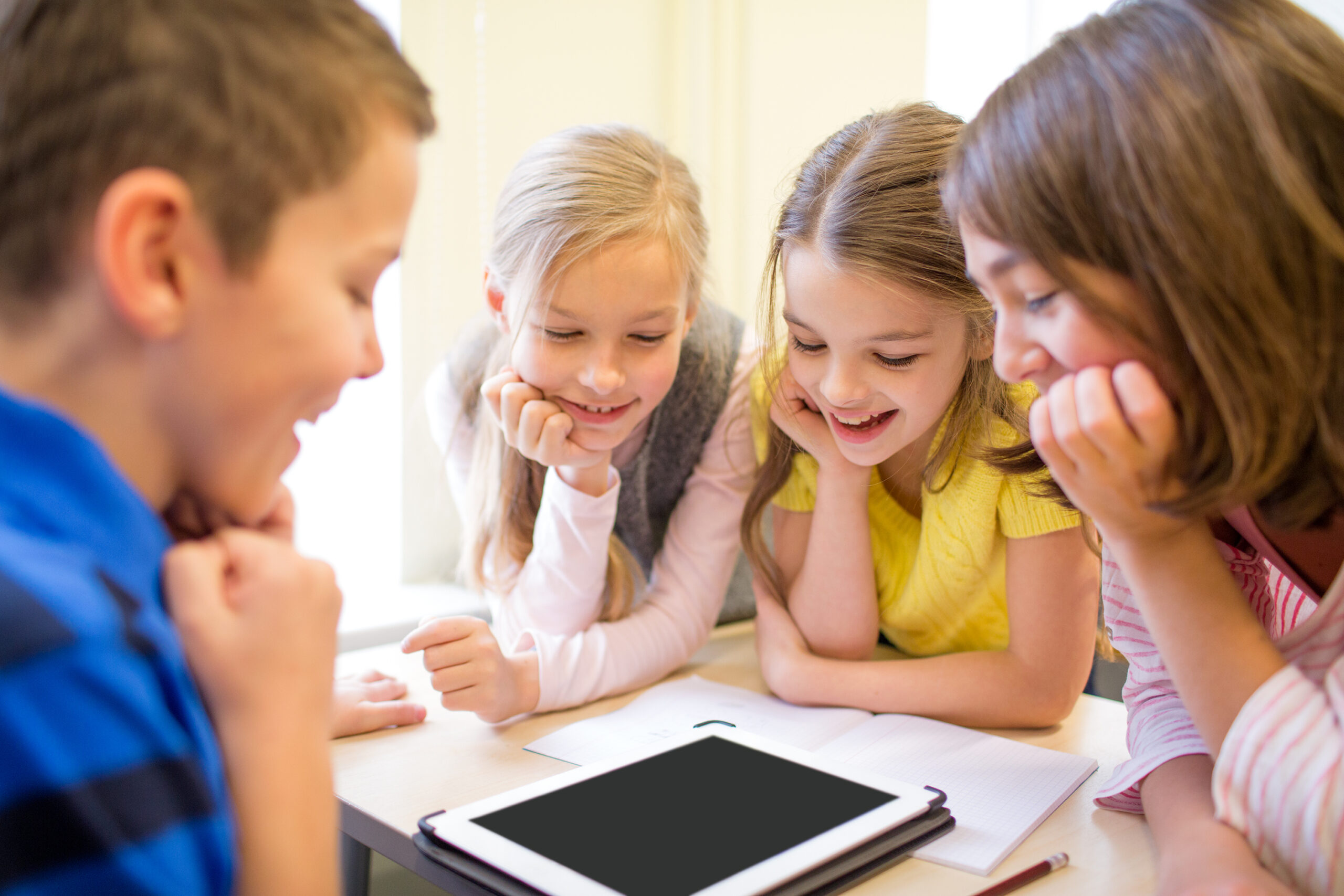 Group of 4 children leaning on a table looking down at a tablet