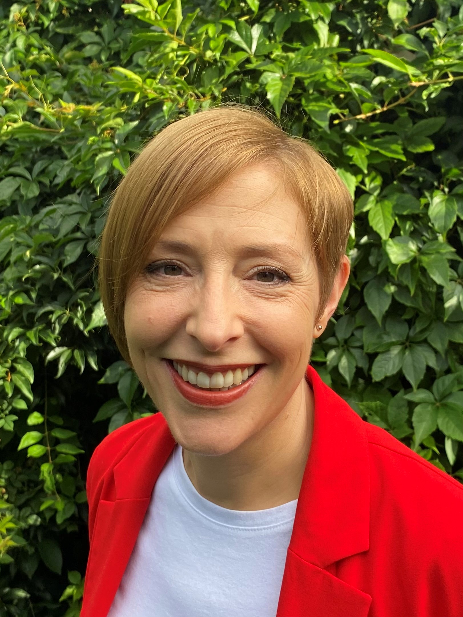 Vanessa is wearing a white top and red jacket, she's smiling at the camera with green foliage behind her